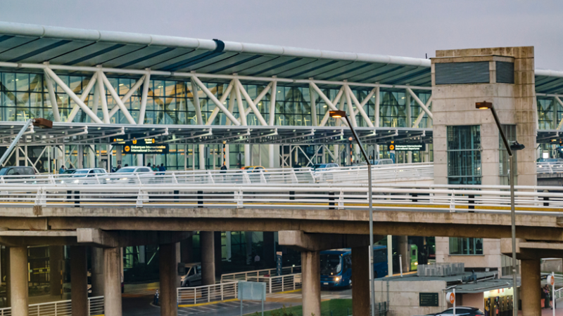 Exterior del aeropuerto internacional de Santiago con pasarela elevada, señales de embarque y buses al atardecer.