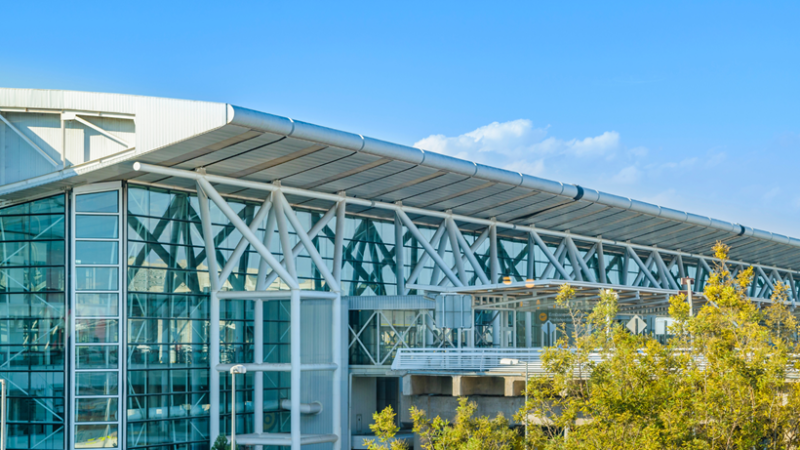 Terminal de aeropuerto moderno con estructura metálica, grandes ventanales y árboles bajo cielo azul despejado.
