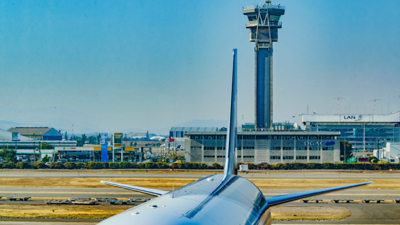 Cola de avión en pista con torre de control y edificio DGAC del aeropuerto internacional de Santiago al fondo.
